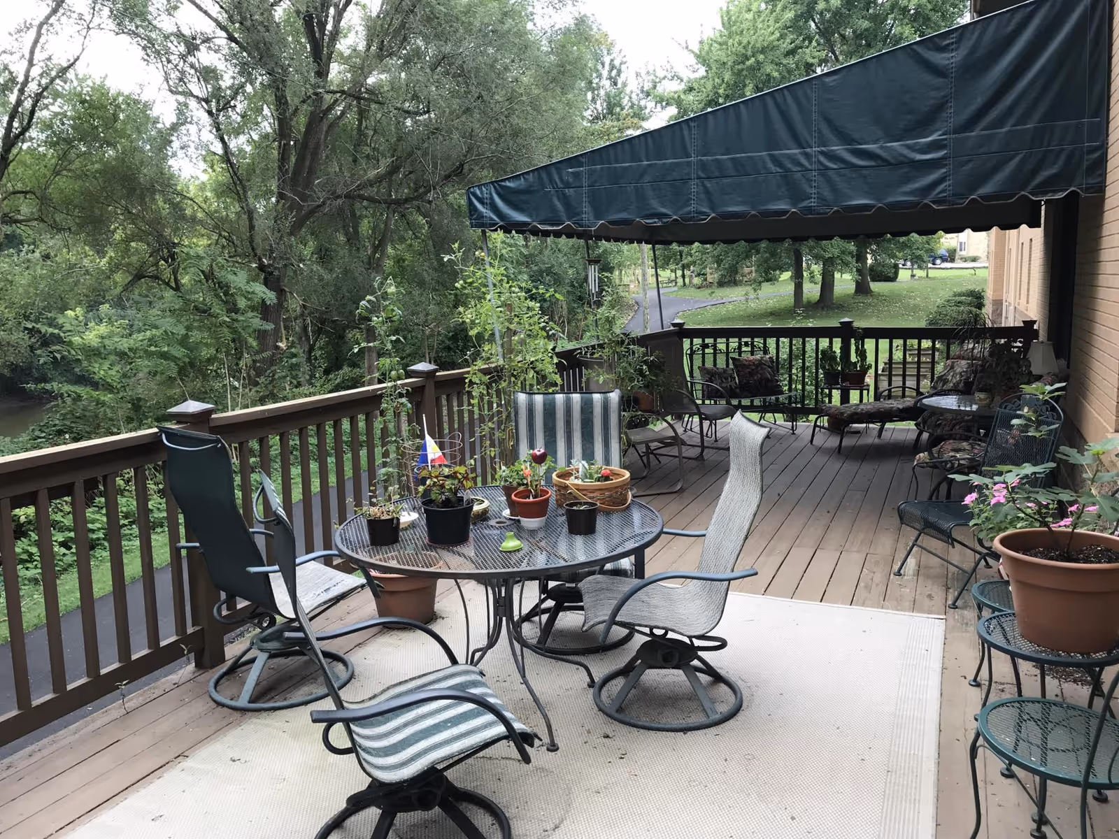 A furnished wooden deck with a round table, several chairs and potted plants under a green awning overlooking a tree-lined yard.