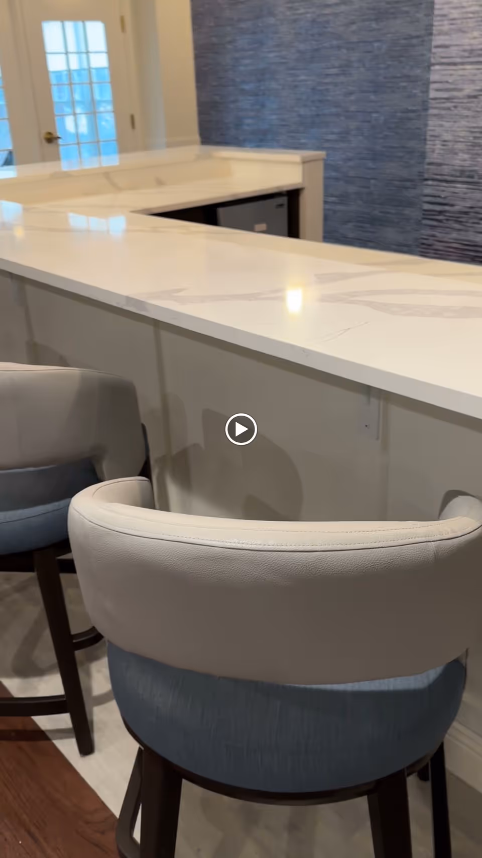 Two cushioned bar stools with light beige backs and blue seats positioned in front of a white marble countertop bar. Behind the bar is a small refrigerator and a textured blue wall. A door with glass panels is visible in the background.