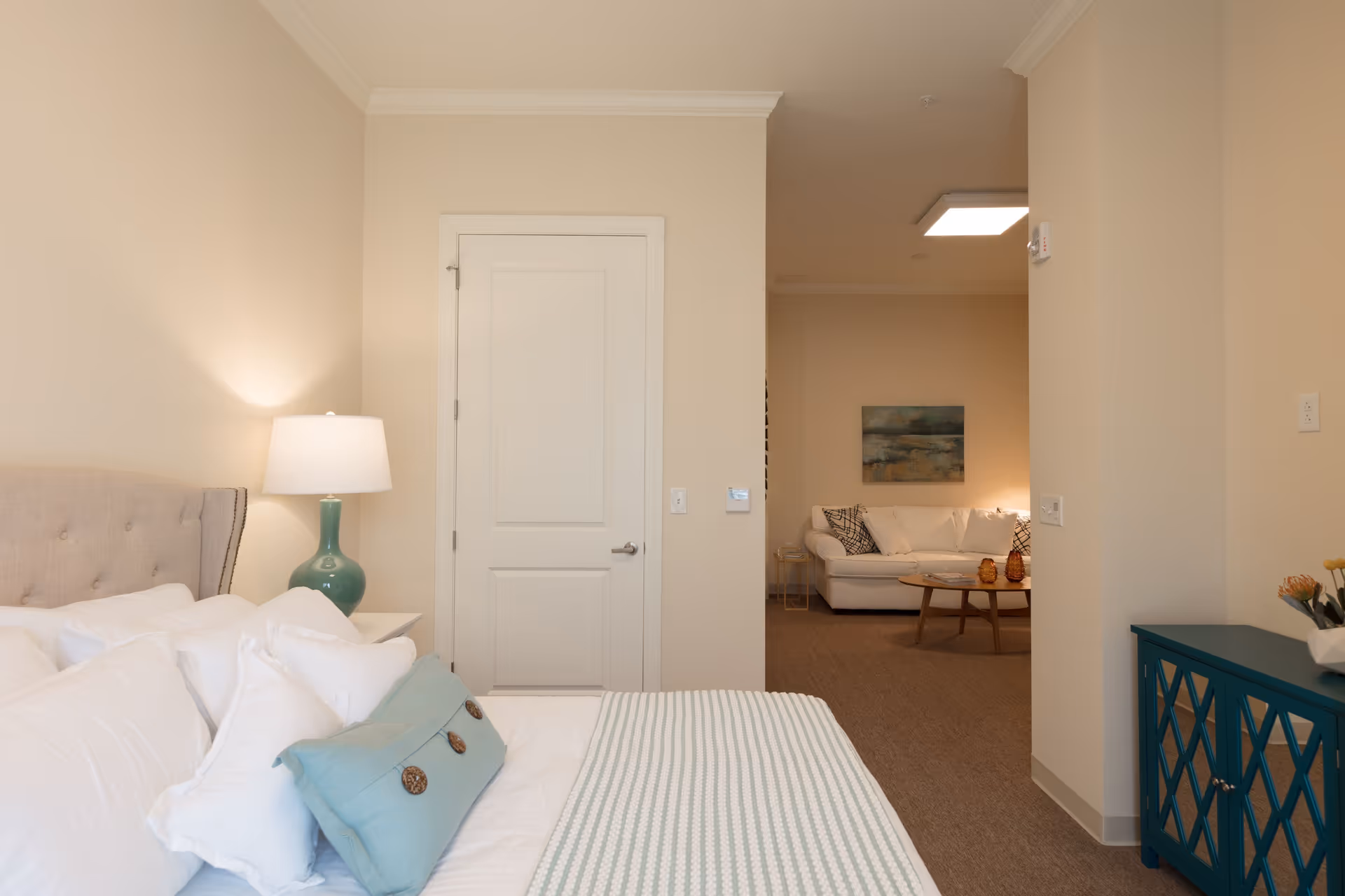 A bedroom with a neatly made bed featuring white pillows and a light blue decorative pillow. A bedside table with a green lamp is next to the bed. The room has beige walls and a white door. Through an open doorway, a living room area is visible with a white couch, a wooden coffee table, and a painting on the wall.