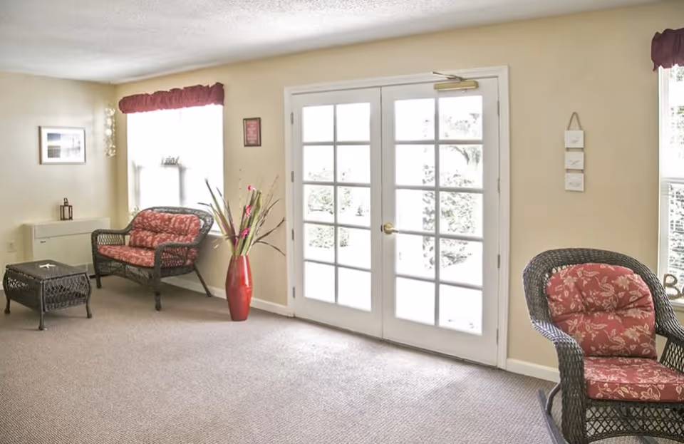 Bright sitting area with wicker chairs and floral cushions, a red vase, and French double doors.