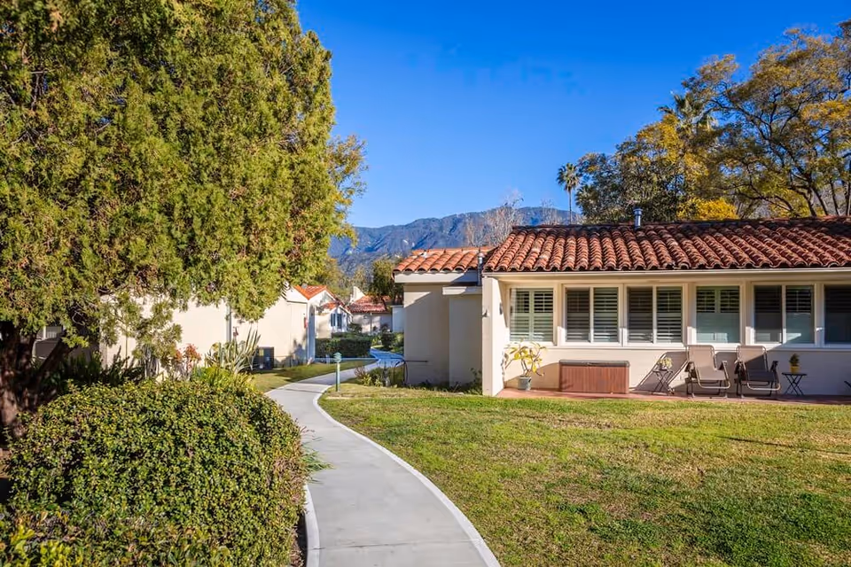 A sunny outdoor scene at Westminster Gardens featuring a curved concrete pathway bordered by green bushes and trees. To the right, there is a single-story building with a red tiled roof, white walls, and several windows with shutters. Patio chairs and a small table are arranged outside the building. Mountains and a clear blue sky are visible in the background.