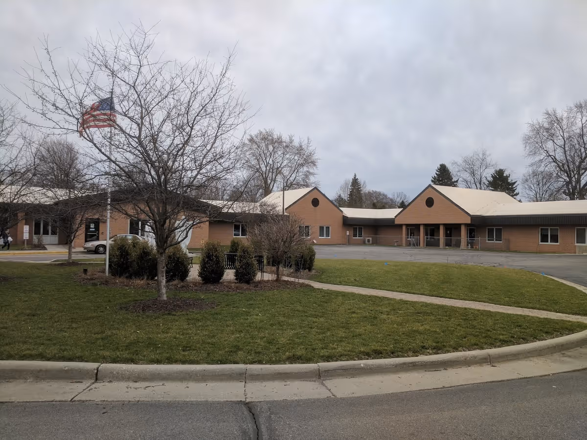 Exterior view of a single-story brick building with a white roof, surrounded by a lawn with some bushes and leafless trees. An American flag is flying on a flagpole near the building. The sky is overcast.