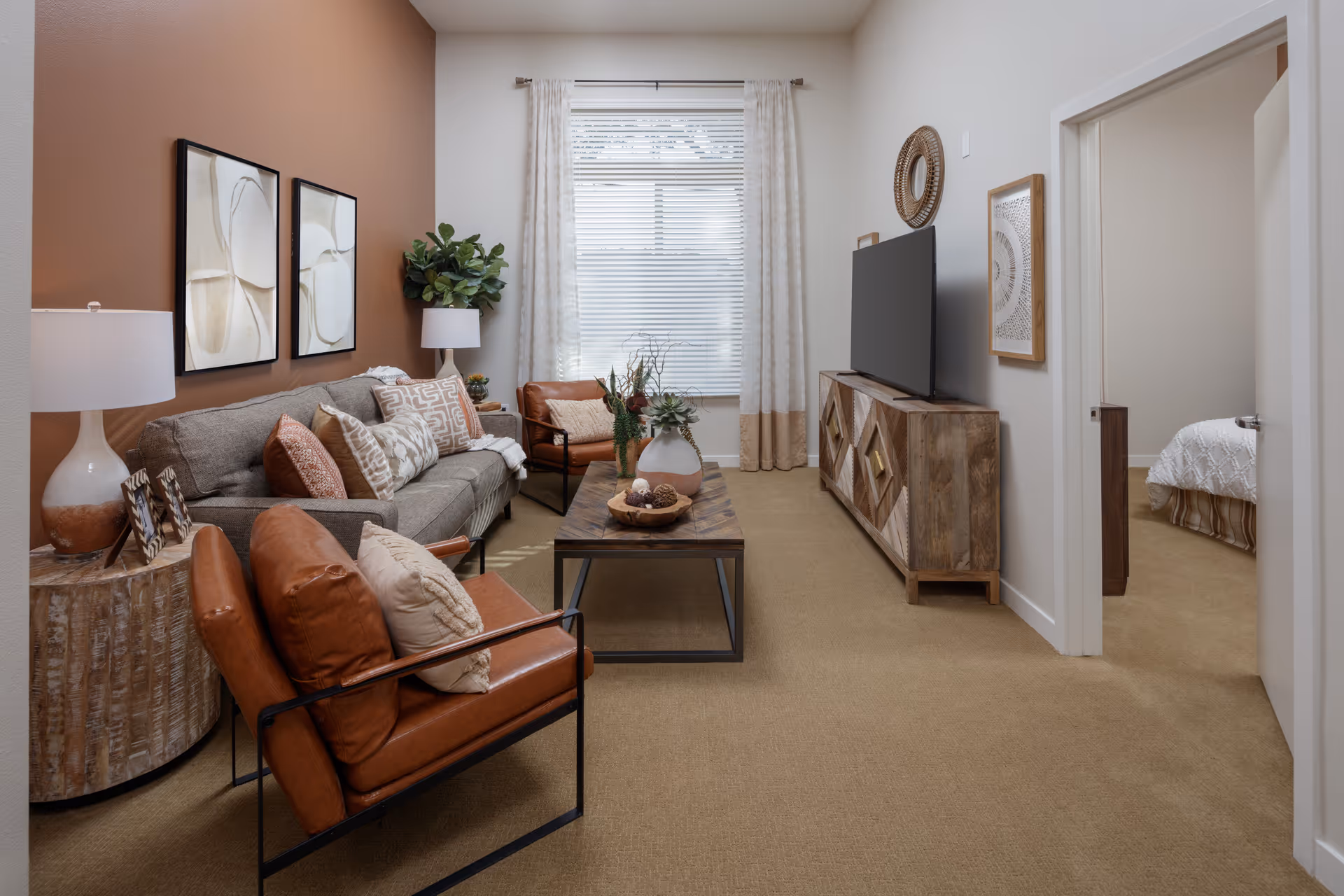 A cozy living room with a gray sofa adorned with multiple patterned cushions, two brown leather armchairs with pillows, a wooden coffee table with decorative items, a wooden TV stand with a flat-screen TV, and a window with white curtains and blinds. The walls are painted white and one accent wall is a warm brown color. A doorway leads to a bedroom with a white bedspread visible.