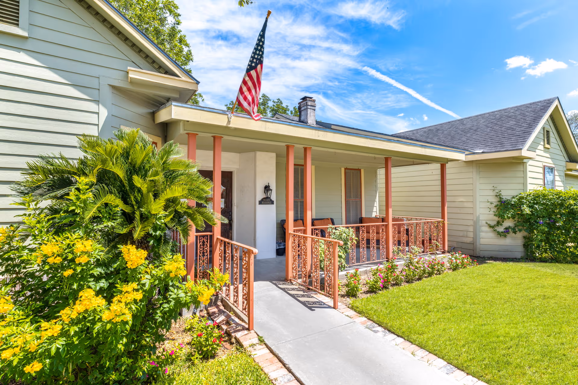 Front exterior view of a light green house with a covered porch supported by orange columns and railings. The porch has seating and an American flag mounted above. There are yellow flowers and green shrubs along the walkway leading to the porch, and a well-maintained lawn on the right side under a bright blue sky with some clouds.