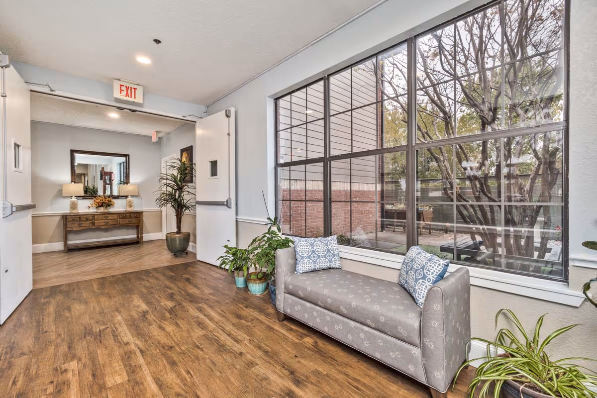 Sunlit senior living seating area with a cushioned bench and pillows by large windows, potted plants, and a console table with a mirror near double doors.
