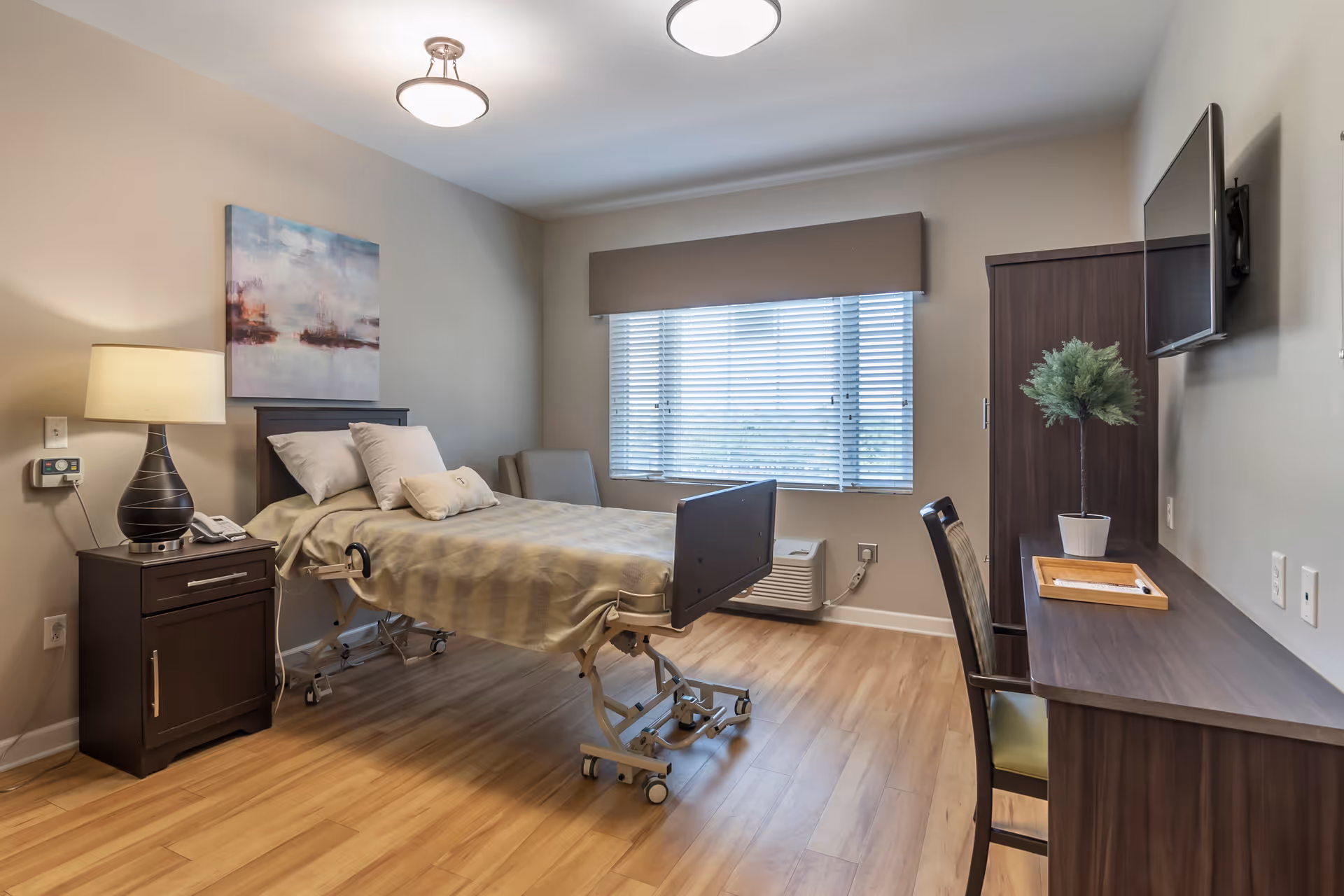 A well-lit, clean bedroom in a senior living facility featuring a hospital-style bed with beige bedding, a nightstand with a lamp and phone, a chair near a window with blinds, a wall-mounted TV, a desk with a chair, and a small potted plant on the desk.