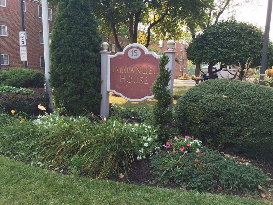 Outdoor garden area with various green shrubs, flowering plants, and a sign that reads 'Immanuel House Retirement Living' with the number 15 and the year 1865. A brick building and a speed limit sign are visible in the background.