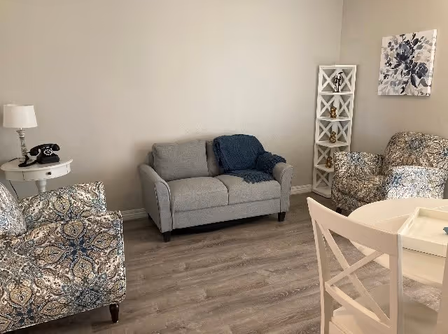 A cozy living room with a gray loveseat, two patterned armchairs, a white round table with chairs, a white side table with a lamp and a vintage black rotary phone, a white corner shelf with decorative items, and a floral wall art on a beige wall. The floor is wood laminate.