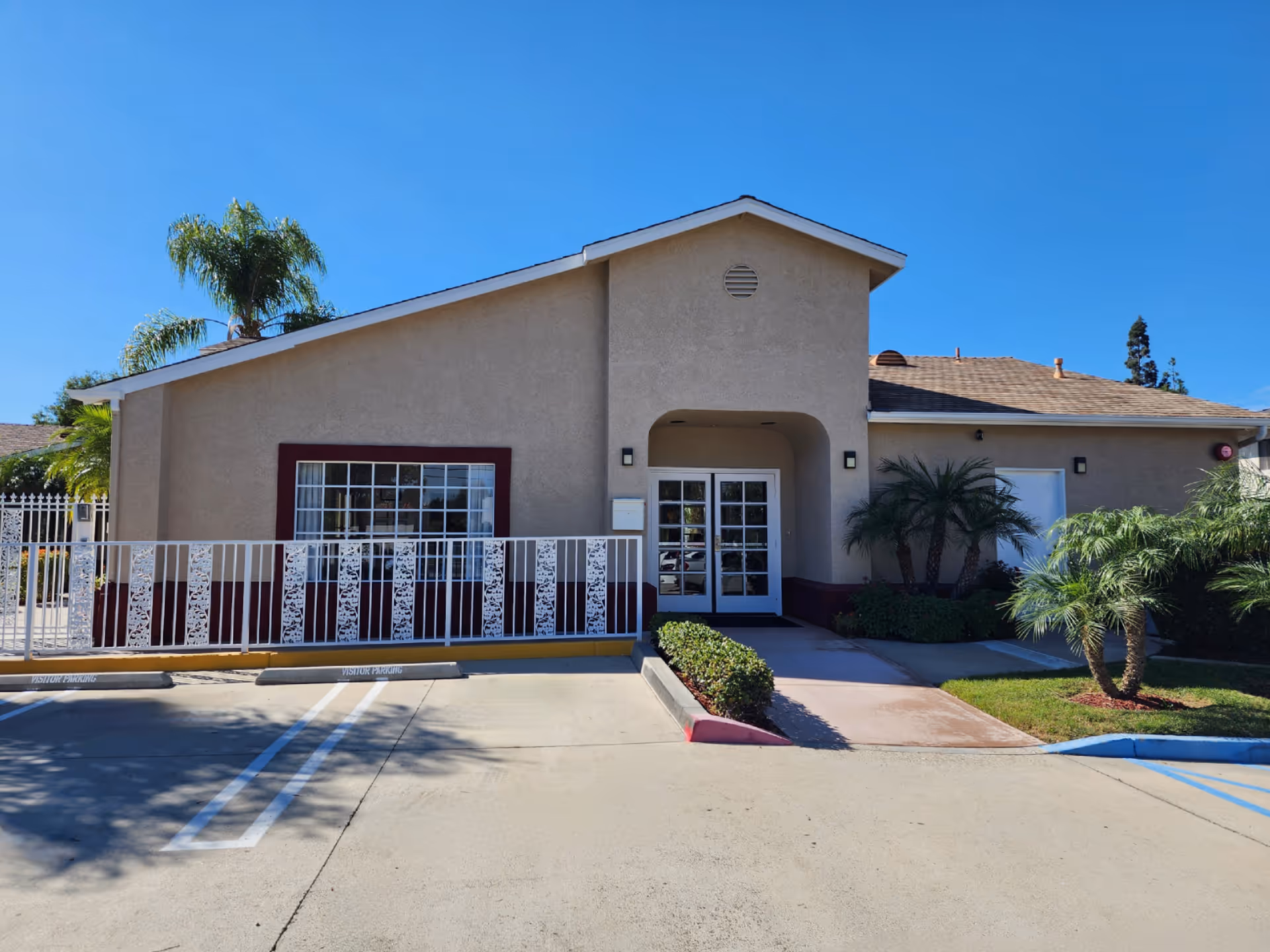 Exterior front view of a single-story building with beige walls and a brown roof under a clear blue sky. The entrance has double glass doors under a small covered porch. There is a white decorative metal fence in front of a large window to the left of the entrance. The parking area in front has marked visitor parking spaces. Palm trees and other greenery are planted around the building.