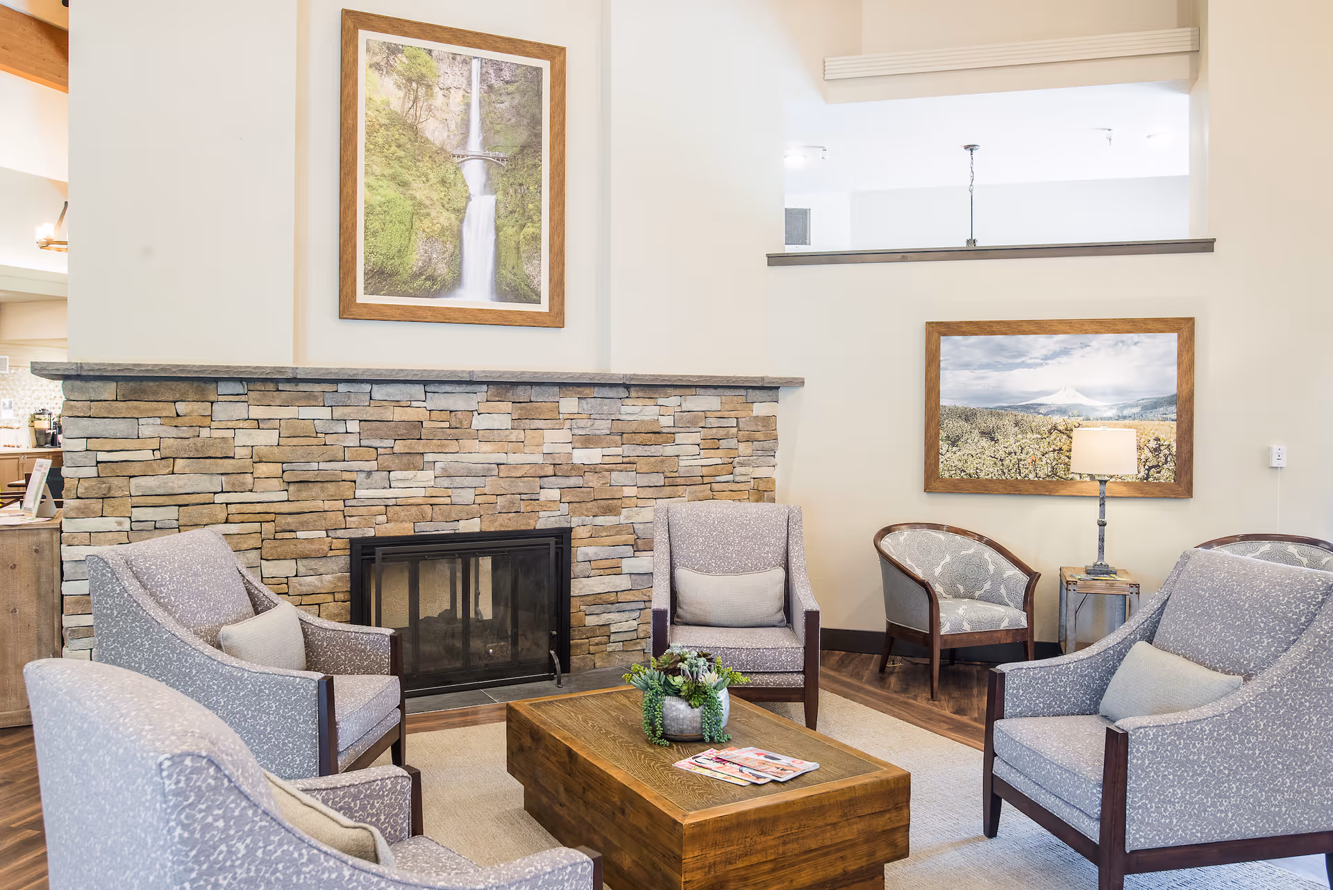 A cozy living room area with four gray upholstered armchairs arranged around a wooden coffee table with a small plant and magazines on it. The room features a stone fireplace with a framed picture of a waterfall above it. Another framed landscape picture and a table lamp are visible on the right side of the room. The walls are light-colored, and the floor has a wood finish.