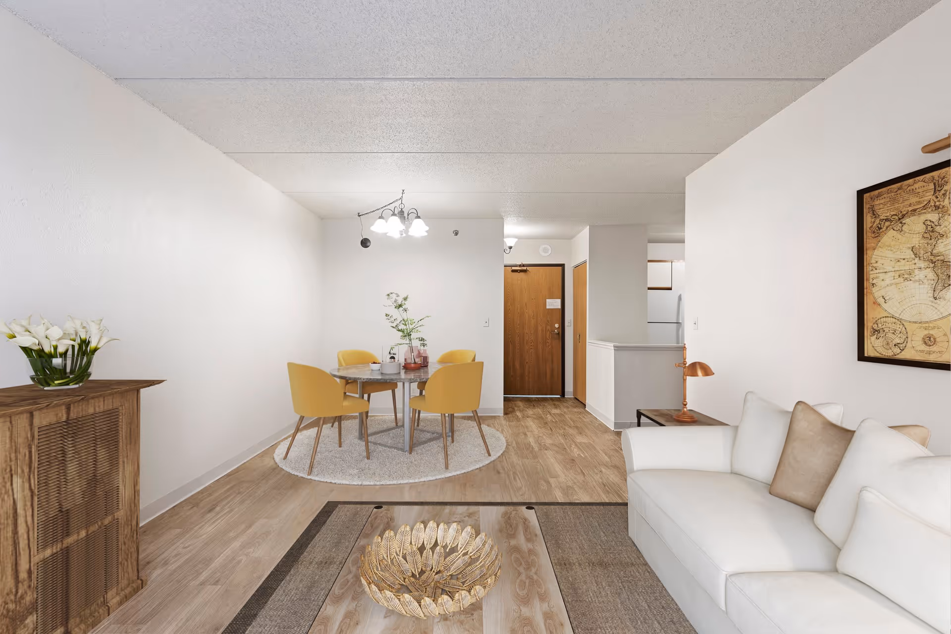 Bright living and dining area with a white sofa, coffee table, round dining table with yellow chairs, and a wooden entry door.