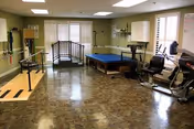 Rehabilitation gym interior with parallel bars, a therapy table, stationary bikes, a small practice bridge and other exercise equipment under ceiling lights.