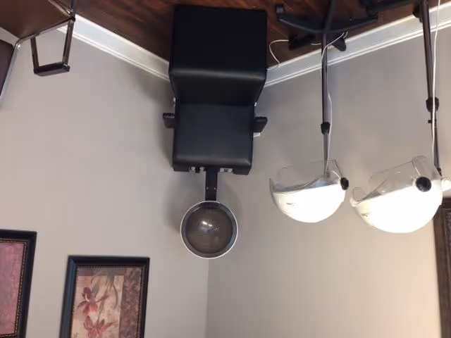 Interior view of a hair salon area with a black salon chair and a hair washing basin attached. Two white hair dryer chairs with hoods are positioned nearby. The walls are light-colored with framed floral artwork hanging on them. The floor is wooden.