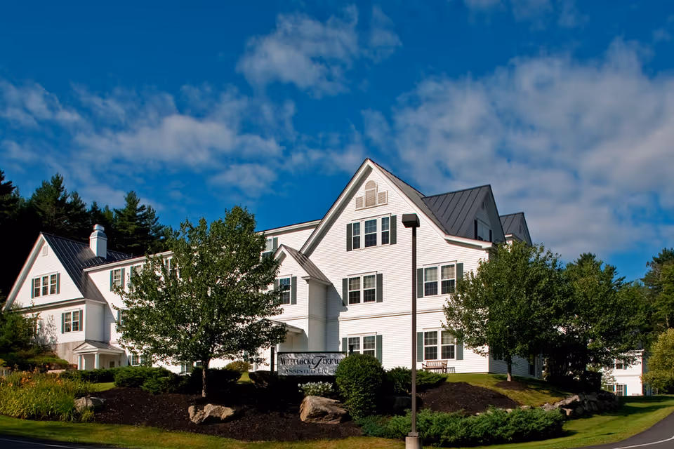 Exterior view of a large white multi-story building with black roof, surrounded by trees and landscaping under a partly cloudy blue sky. A sign in front reads 'Wheelock Terrace Assisted Living'.