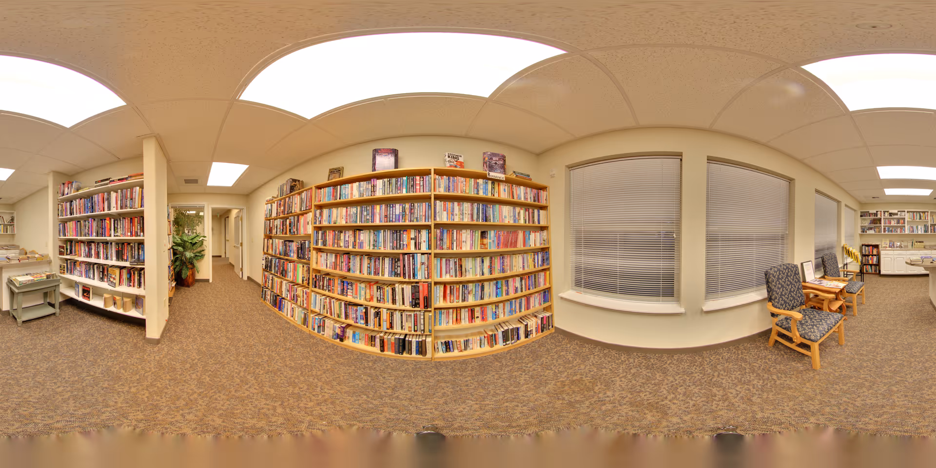 Interior view of a senior living facility library with bookshelves filled with books along the walls, carpeted floor, ceiling lights, two chairs with patterned upholstery near windows with blinds, and a small table with a computer in the background.