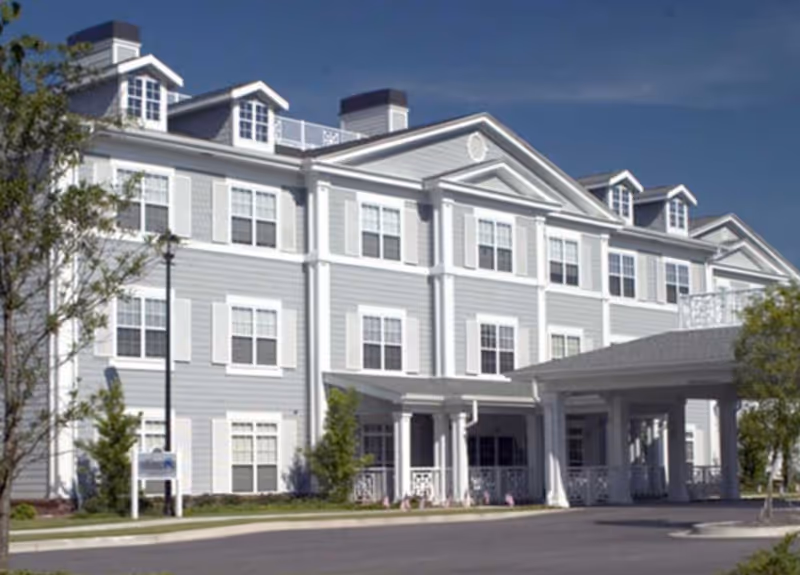 Exterior view of a large, three-story senior living facility building with light gray siding, white trim, multiple windows, and a covered entrance. There are small trees and a clear blue sky in the background.