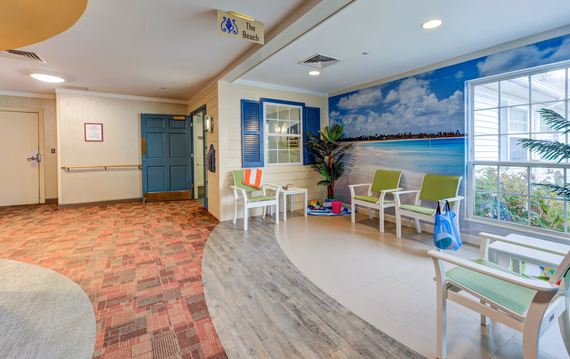 A bright indoor seating area with green cushioned chairs and white frames arranged around a small white table. The wall features a large mural of a beach scene with blue skies and ocean waves. There are potted plants and a window letting in natural light. The floor transitions from wood-like flooring to tile and carpet. A sign above reads 'The Beach'.
