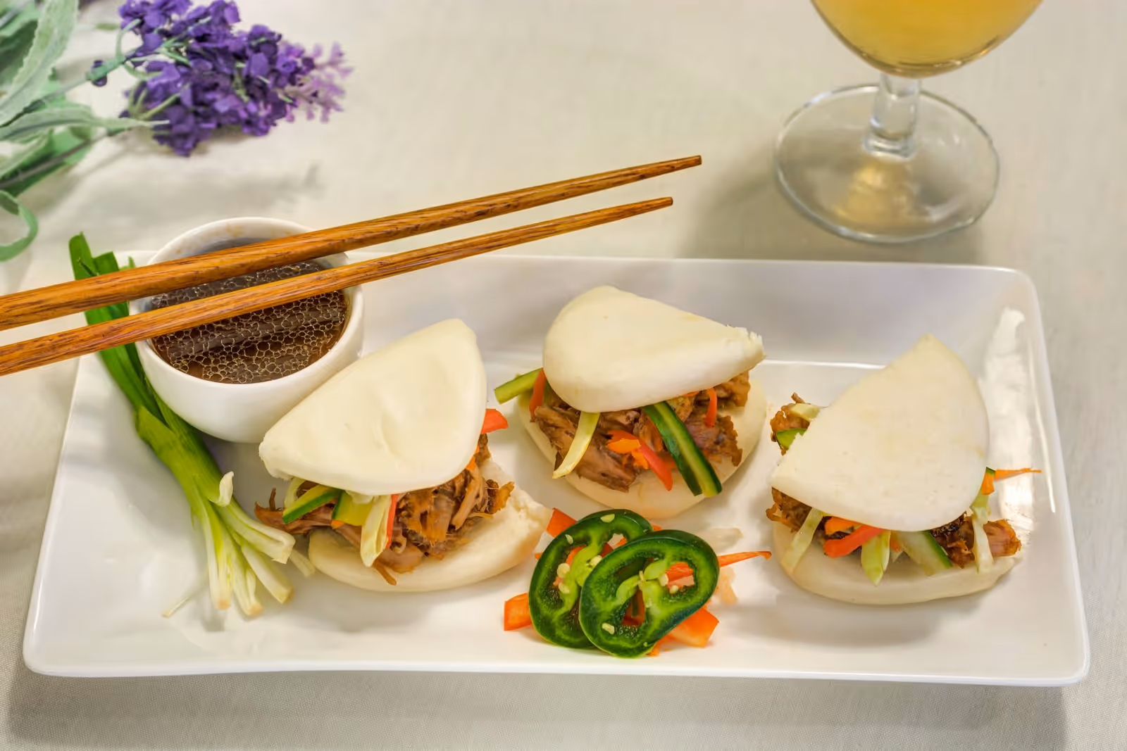 Three steamed bao buns filled with shredded meat and vegetables on a rectangular white plate, accompanied by sliced jalapeños, a small bowl of dipping sauce with chopsticks resting on it, green onions, and a glass of light-colored beverage in the background.