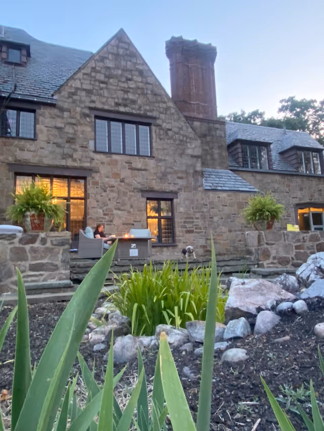 Stone building with a large chimney and multiple windows at dusk. In front of the building, there is a stone patio with outdoor seating where a person is sitting. There are plants and rocks in the foreground, and two large potted ferns on stone pillars flanking the patio.