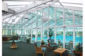 Glass-enclosed indoor pool area with seating, tables, and potted plants under a white-framed atrium roof.