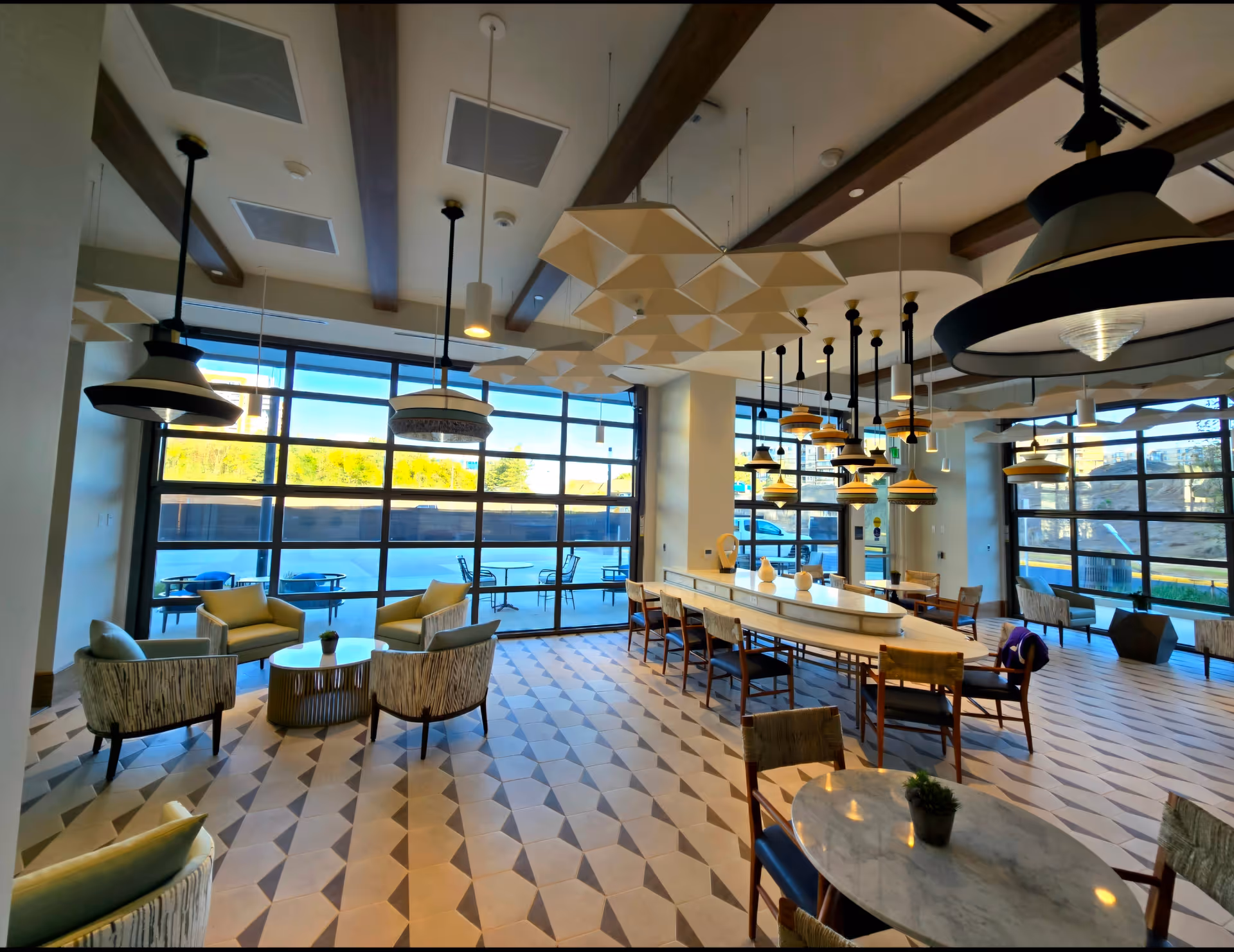 Spacious bright communal dining and lounge area with floor-to-ceiling windows, pendant lights, a long central table and multiple seating areas.