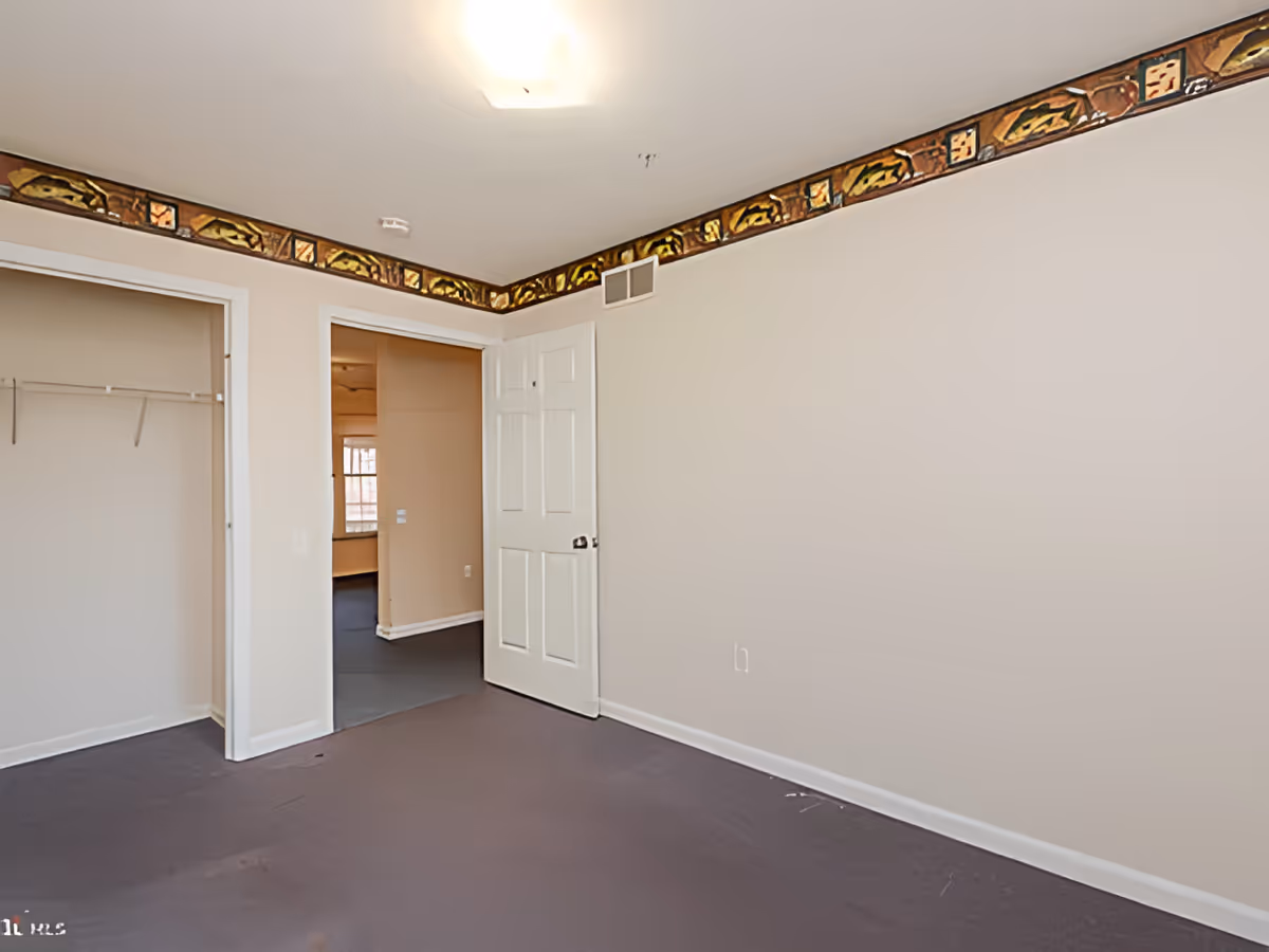 Empty room with beige walls, a white door slightly open leading to another room, an open closet with a hanging rod, and a decorative border near the ceiling featuring a pattern with fish and geometric shapes.