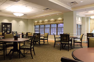 A spacious dining room with multiple round tables and chairs arranged neatly. The room has large windows with blinds, soft lighting from ceiling fixtures, and a cabinet with decorative items along one wall.