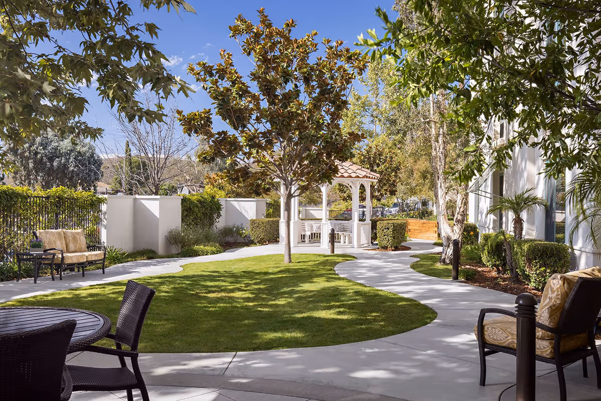 Outdoor garden area with a curved concrete pathway, green grass, trees, and shrubs. There are several seating areas including cushioned benches and chairs around tables. A white gazebo with a tiled roof is visible in the background under a clear blue sky.