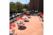 Outdoor patio area with several round tables and chairs, each table shaded by a red umbrella. The patio overlooks a parking area with cars and trees in the background, along with a tall building nearby.