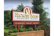 Outdoor view of a sign for Hickory House Nursing Home with the words Outpatient Therapy Services below it, surrounded by greenery and trees.