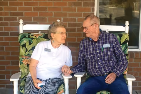 An elderly woman and an elderly man sitting on cushioned rocking chairs outside against a brick wall. They are holding hands and looking at each other with smiles.