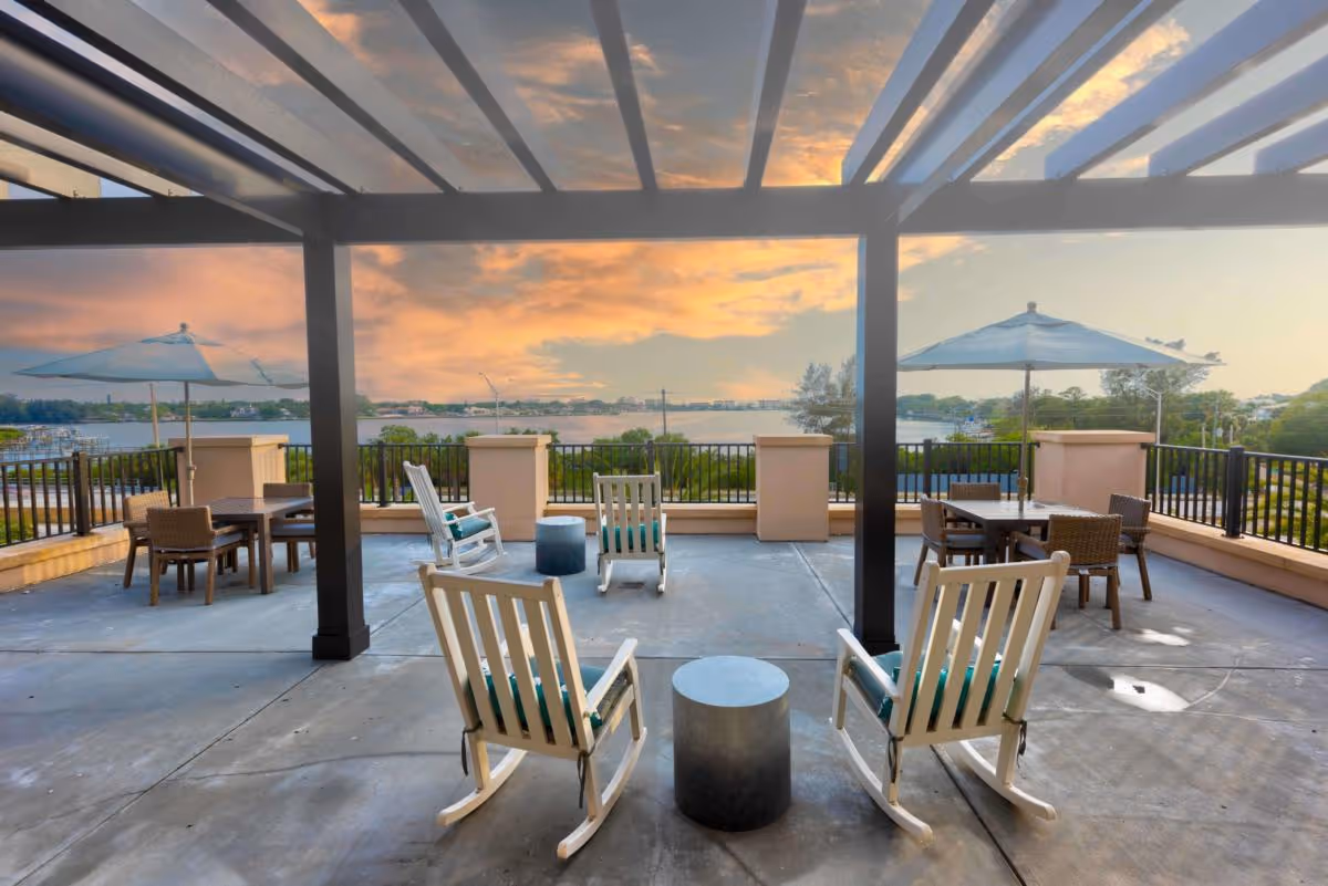 Rooftop patio with white rocking chairs, tables and umbrellas under a pergola overlooking a waterfront at sunset.