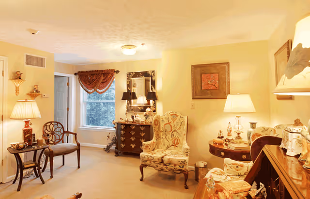Cozy living room with two upholstered chairs, a small table, decorative lamps, and a window with curtains.