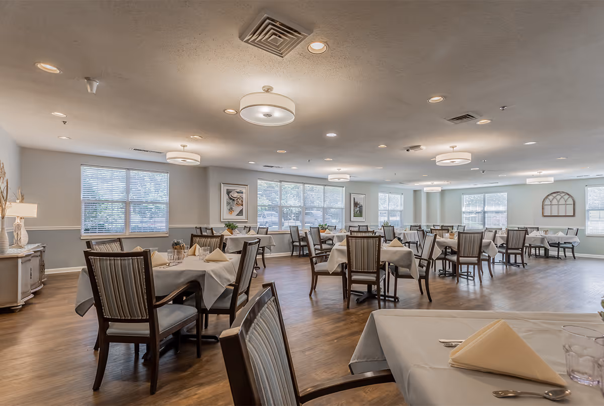 A spacious dining room with multiple tables covered in white tablecloths, each set with folded napkins, glasses, and silverware. The room has wooden flooring, several large windows letting in natural light, and modern ceiling lights. The walls are decorated with framed artwork and a decorative window frame.