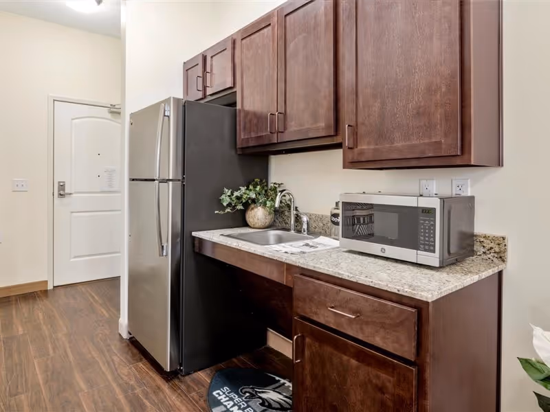 A small kitchen area featuring dark wood cabinets, a granite countertop with a stainless steel sink, a microwave, and a stainless steel refrigerator. The floor is wooden, and there is a white door in the background.