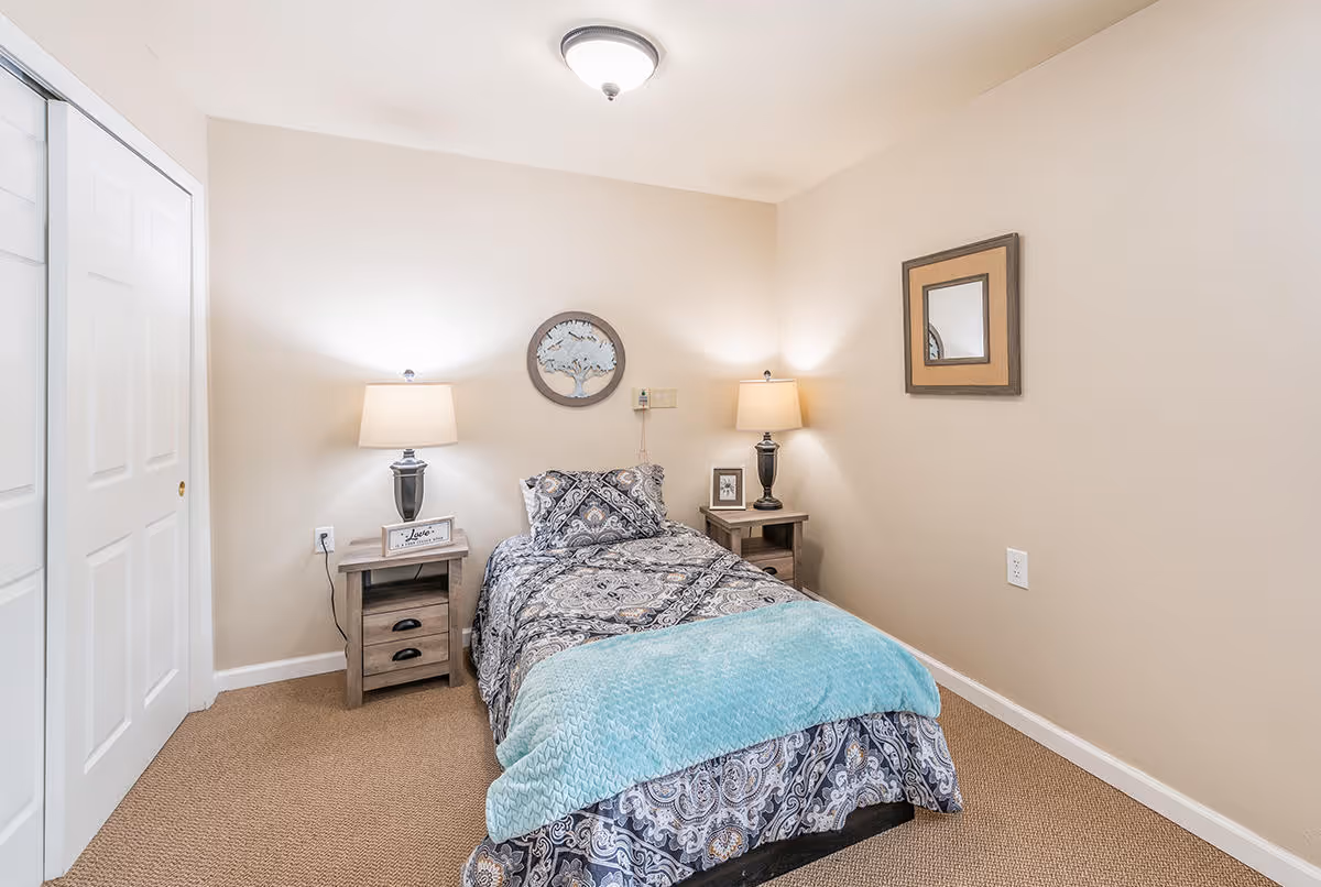 A small bedroom with a single bed covered in patterned bedding and a light blue throw blanket. There are two wooden nightstands on either side of the bed, each with a lamp. The walls are beige, with a round decorative piece above the bed and a square mirror on the adjacent wall. A white closet door is visible on the left side of the room.
