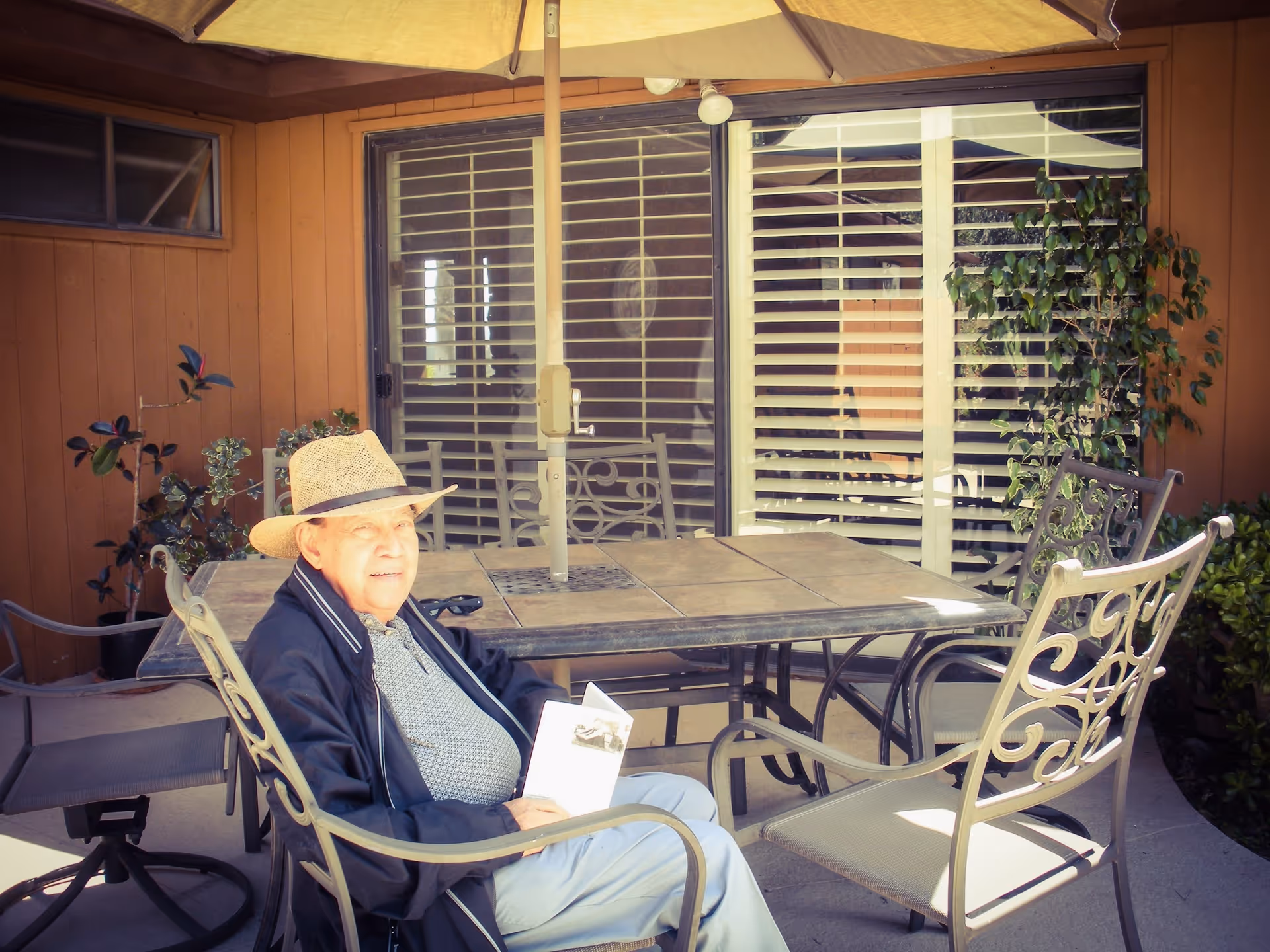 An elderly man wearing a straw hat and dark jacket is sitting on a metal patio chair at an outdoor table with a large umbrella. He is holding a piece of paper and smiling. The setting is a patio area with brown wooden walls, potted plants, and sliding glass doors with white blinds.