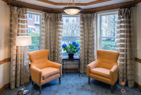 Two upholstered armchairs and a floor lamp arranged around a small table with a potted plant in front of three large windows with striped curtains.