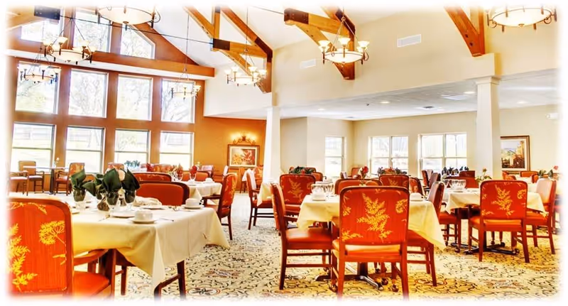 Bright dining room with multiple tables set with white tablecloths and red upholstered chairs under a wooden‑beamed vaulted ceiling and large windows.