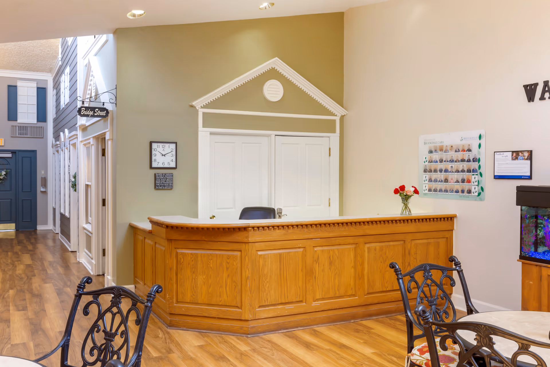 Reception area with a wooden front desk, a clock on the wall, a small vase with red and white flowers on the desk, and a hallway with doors and a sign that reads 'Bridge Street'. There are also decorative chairs and tables in the foreground and a fish tank on the right side.