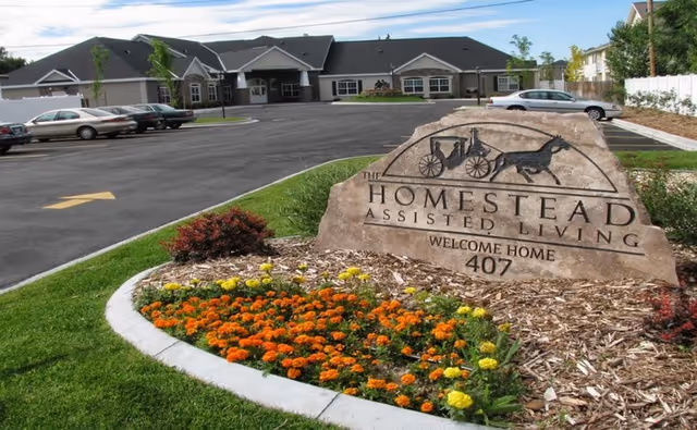 Exterior view of Homestead Assisted Living facility with a large stone sign in the foreground that reads 'The Homestead Assisted Living Welcome Home 407'. The sign is surrounded by a flower bed with orange and yellow flowers. The building and parking lot with several parked cars are visible in the background under a partly cloudy sky.