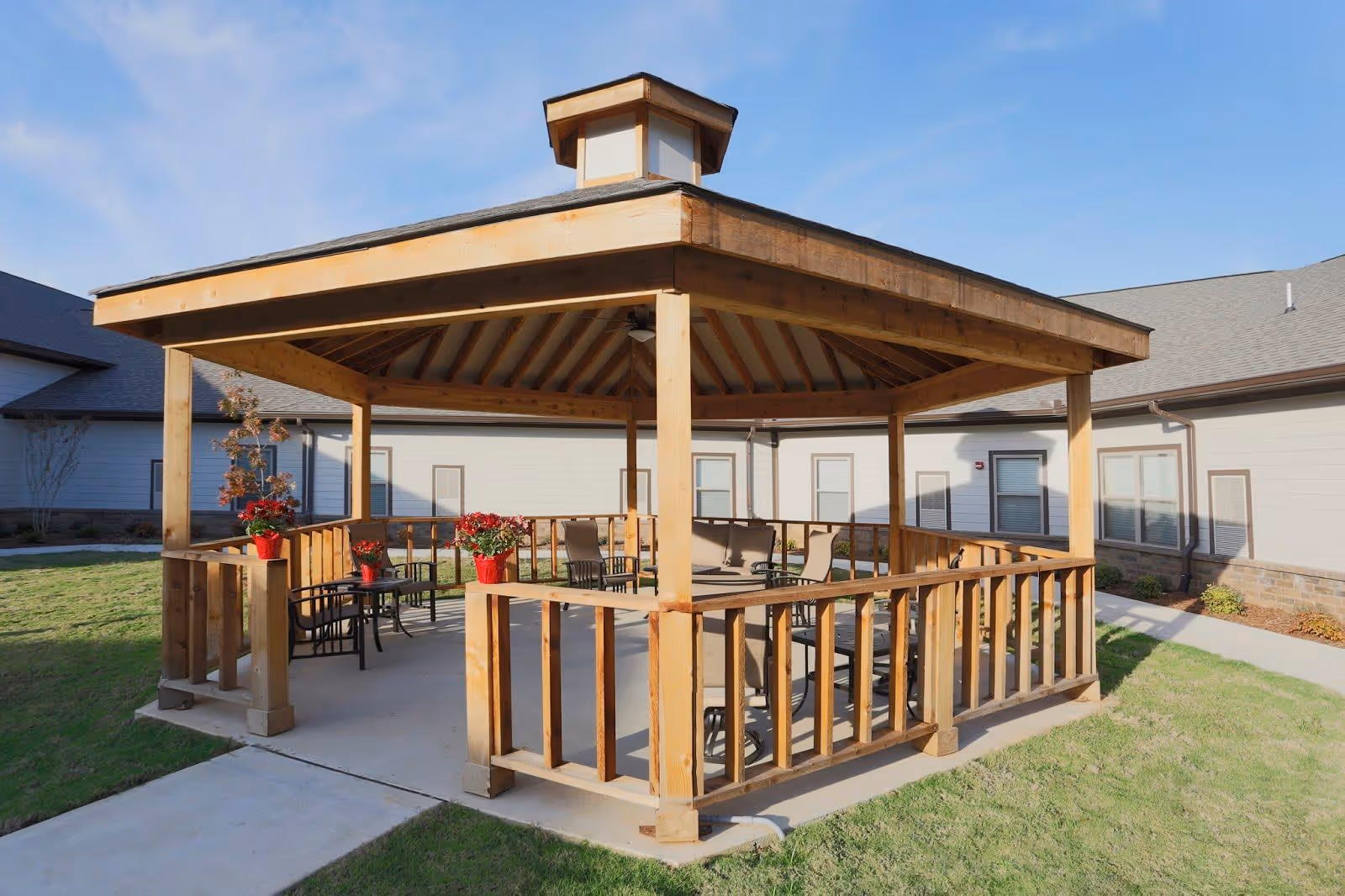 A wooden gazebo with a pitched roof situated on a concrete patio surrounded by grass. Inside the gazebo are several chairs and tables, along with red flower pots placed on the railing. The background shows a single-story building with beige siding and multiple windows under a clear blue sky.