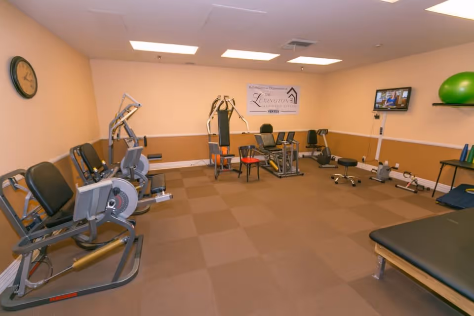Exercise room in Lexington Assisted Living facility with various fitness equipment including leg press machines, a multi-gym machine, stationary bikes, a wall-mounted TV, and exercise balls. The room has beige walls with a brown lower half, a clock on the wall, and a sign for Lexington Assisted Living on the back wall.