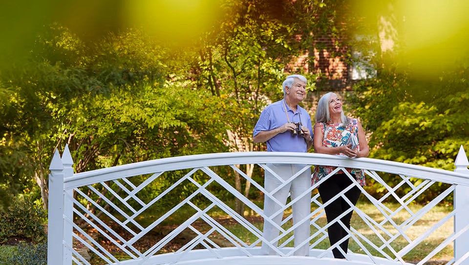 An elderly man and woman standing on a white decorative bridge in a lush green garden. The man is holding binoculars and the woman is holding a book, both looking upwards and smiling.
