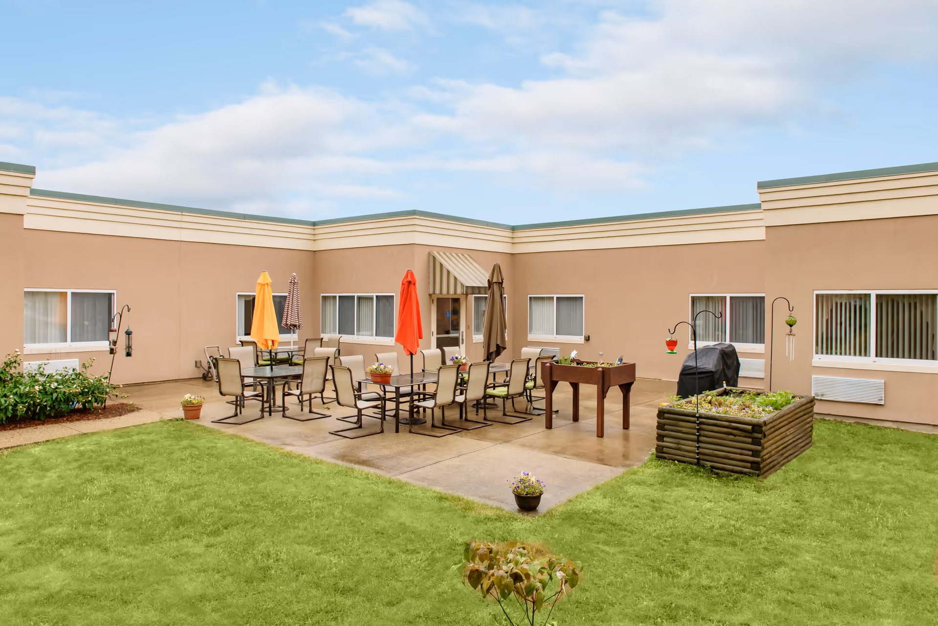Outdoor courtyard area with green grass, patio tables and chairs with colorful umbrellas, a foosball table, a barbecue grill, and a raised garden bed, surrounded by beige building walls with windows.