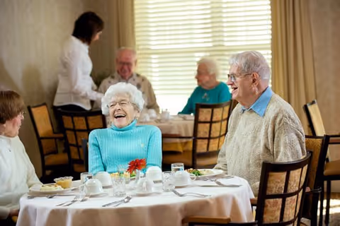 Elderly people sitting at round dining tables in a well-lit room, smiling and engaging in conversation. A woman in a turquoise sweater is laughing, and a man in a beige sweater is smiling at her. Other seniors and a staff member are visible in the background.