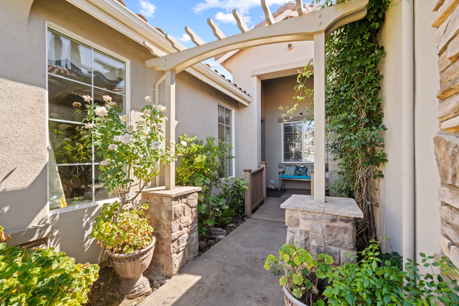 Outdoor walkway with stone pillars and a pergola leading to a covered porch area with a bench and cushions. The walkway is flanked by potted plants and greenery, with windows on the left side of the building.