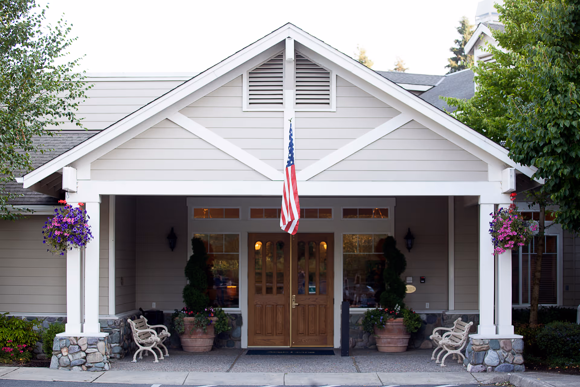 Covered front entrance of Aegis Living Callahan House with double wooden doors under a gabled portico, an American flag, hanging flower baskets and benches.