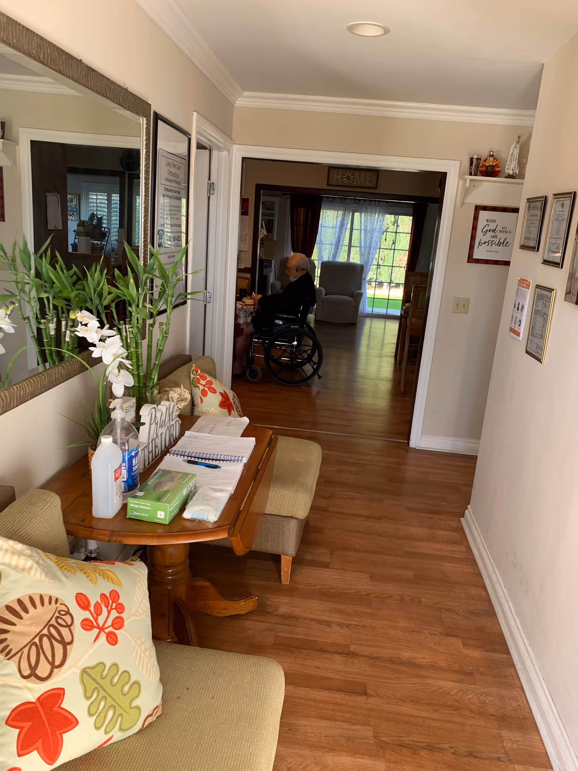Interior hallway and seating area in a senior living facility with a small table holding supplies and a person in a wheelchair visible in the adjoining room.