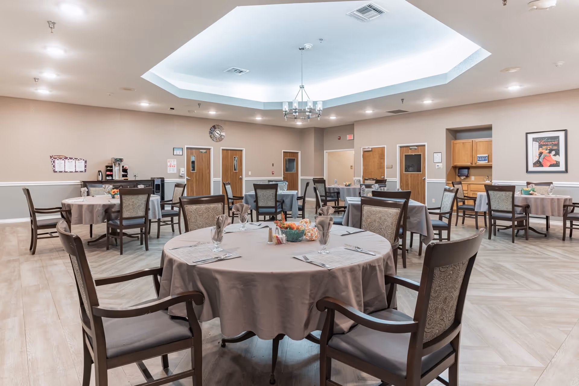 A spacious dining room with multiple round tables covered with beige tablecloths, each set with napkins, glasses, and silverware. The room has wooden chairs with cushioned seats and backs, light-colored wood flooring, beige walls, and a large octagonal recessed ceiling with a modern chandelier. There are several wooden doors along the walls, a coffee station, and framed artwork on the walls.
